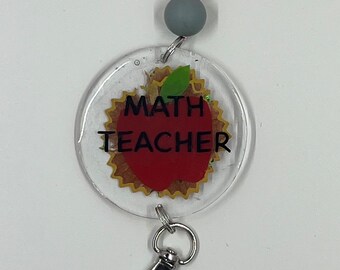 Puede incluir: Un llavero redondo transparente con un gráfico de manzana roja y el texto "MATH TEACHER". El llavero tiene un clip plateado.