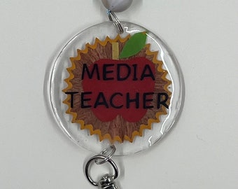 Puede incluir: Un portagafete de plástico transparente con un gráfico de manzana roja y el texto "MEDIA TEACHER". El portagafete tiene un clip giratorio plateado.