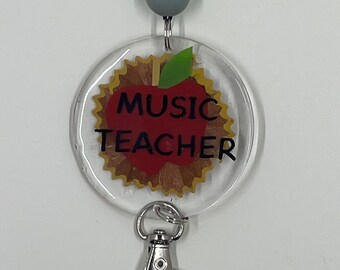 Puede incluir: Un portagafete de plástico transparente con un diseño de manzana roja y el texto "MUSIC TEACHER". El portagafete tiene un clip plateado.
