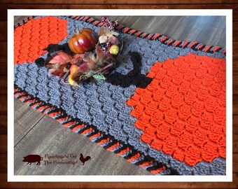Puede incluir: Un camino de mesa tejido a crochet con un esquema de color gris y naranja. El camino de mesa presenta dos formas de calabaza en naranja con contornos negros. El camino de mesa tiene un borde negro y naranja. El texto "Handmade On The Homestead" es visible en la esquina inferior izquierda de la imagen.