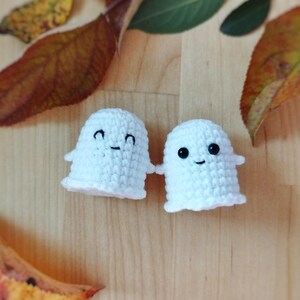 May include: Two small, handmade white crochet ghosts with black eyes and smiles. The ghosts have a simple design with a rounded body and a ruffled bottom edge. The background is a wooden surface with autumn leaves.
