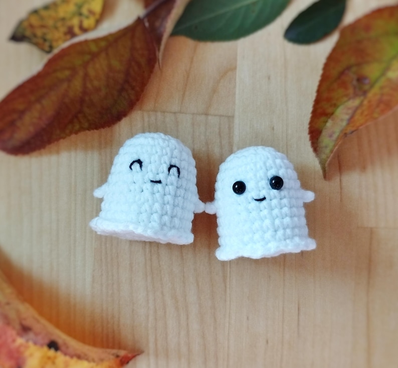 May include: Two small, handmade white crochet ghosts with black eyes and smiles. The ghosts have a simple design with a rounded body and a ruffled bottom edge. The background is a wooden surface with autumn leaves.