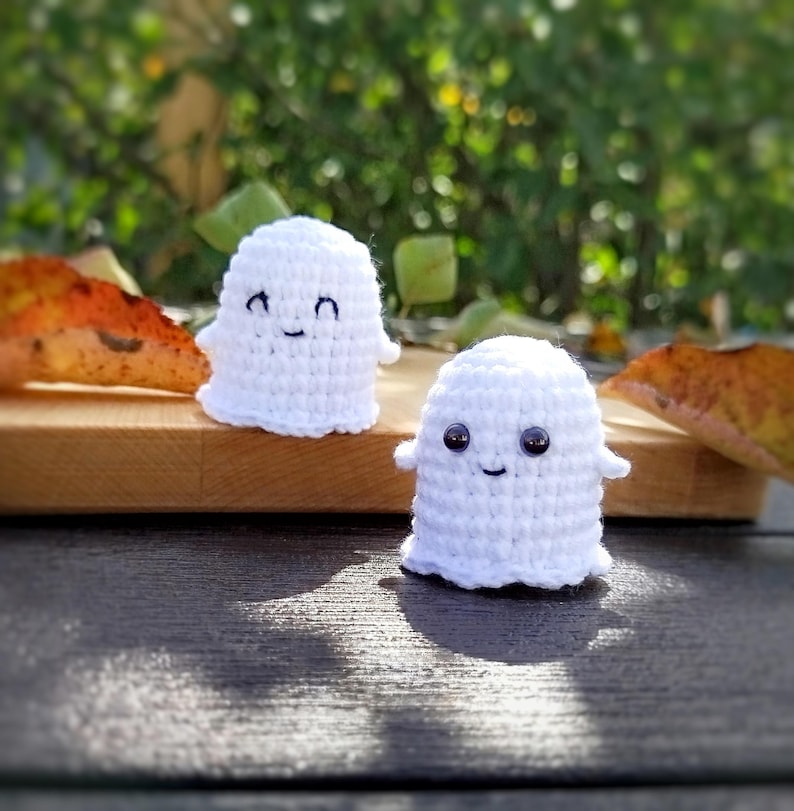 May include: Two small, handmade white crochet ghosts with smiling faces. One ghost has black eyes, the other has black stitched eyes. They are on a wooden surface, with a blurred green and brown background.