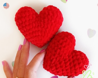 May include: Two plush, red, heart-shaped crochet items. The text "PDF PATTERN" is at the top left, with a small American flag icon. The hearts are held by a person's hand. Decorative heart-shaped confetti is scattered around the hearts.