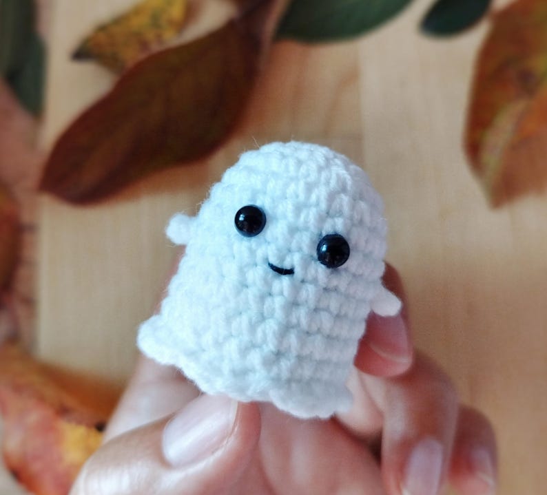 May include: A small, white crocheted ghost with black bead eyes and a stitched smile. The handmade ghost has small, rounded arms and a rounded bottom. The background includes brown and yellow autumn leaves.