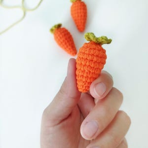 May include: Three small crocheted carrots, one orange carrot is being held in a hand, the other two carrots are on a white surface.
