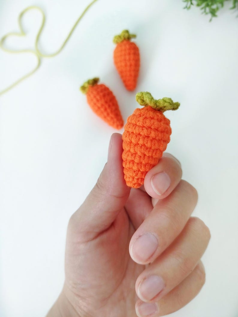 May include: Three small crocheted carrots, one orange carrot is being held in a hand, the other two carrots are on a white surface.