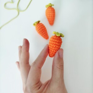 May include: Three crocheted carrots in orange and green. The carrots are small and are arranged on a white surface.