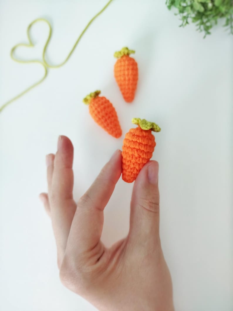 May include: Three crocheted carrots in orange and green. The carrots are small and are arranged on a white surface.