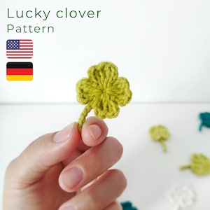 May include: A handmade, green crocheted four-leaf clover is held in a hand. The clover has a stem and is surrounded by other crocheted clovers. The text "Lucky clover Pattern" is at the top of the image, along with the American and German flags.