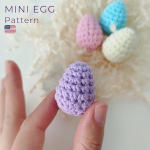 Op de afbeelding: Een close-up van een gehaakt paars ei. Het ei wordt vastgehouden door een hand. De afbeelding toont ook andere gehaakte eieren in roze, blauw en wit.