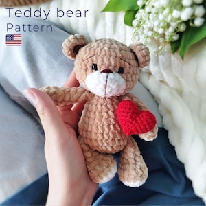 May include: A crocheted brown teddy bear holding a red heart. The bear has a white muzzle and a stitched nose. The text "Teddy bear Pattern" is at the top left with a small American flag. The bear is held in a person's hand.