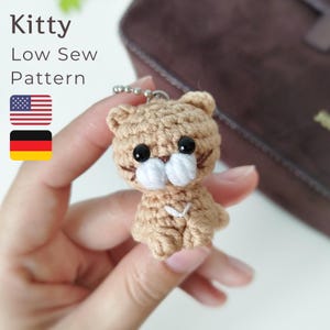 May include: A crocheted tan cat keychain with black eyes and white muzzle. The keychain has a silver chain and a small white V-shaped detail on its chest. The image also includes the text "Kitty Low Sew Pattern" and the American flag.