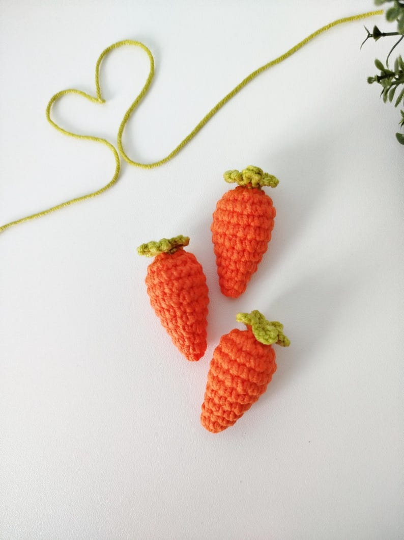 May include: Three crocheted carrots with green tops. The carrots are orange and the tops are green. The carrots are arranged in a triangular shape on a white background.
