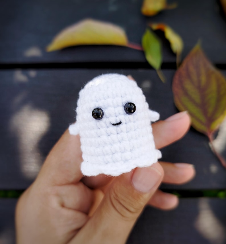 May include: A small, handmade white crochet ghost with black button eyes and a smiling mouth. The ghost has a simple, rounded shape and small arms. The background includes fall leaves on a dark wooden surface.