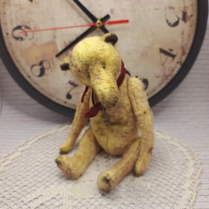 Puede incluir: Un oso de peluche de estilo vintage, principalmente amarillo con sombras más oscuras, sentado sobre un mantel de encaje. Tiene una cinta roja y un colgante. El fondo incluye un reloj grande.