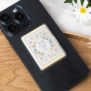 Puede incluir: Un teléfono inteligente negro con un agarre rectangular para teléfono adjunto. El agarre del teléfono tiene un fondo color crema con diseños florales y el texto "HOW LUCKY ARE WE". Las margaritas están en el fondo.