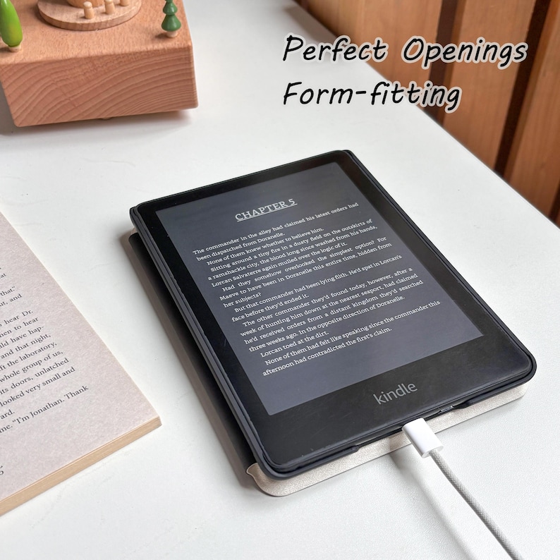 May include: A black Kindle e-reader with a white charging cable, displaying text from Chapter 5. The device is in a light-colored case, with the word "Kindle" visible. The image also includes the text "Perfect Openings" and "Form-fitting".