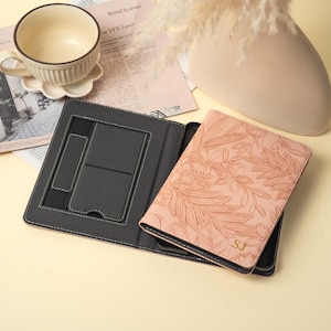 Puede incluir: Una funda negra para tableta con una cubierta en relieve floral rosa claro. La funda está abierta, revelando un interior negro con un soporte para tableta y un soporte. Las iniciales "SJ" están grabadas en dorado en la cubierta.