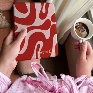 Könnte beinhalten: Eine rot-beige gemusterte Tablethülle mit dem Text "Sweet Em" in goldfarbenen Buchstaben. Die Hülle wird in einer Hand gehalten, mit einer Tasse Kaffee in der anderen Hand. Die Hülle hat ein Retro-Wellen-Design.