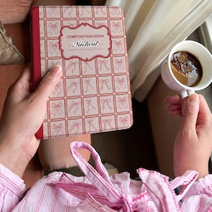 Puede incluir: Un cuaderno de composición rojo y blanco con un patrón de lazo y el nombre "Micheal" en la portada. El libro se sostiene junto a una taza de café. La persona lleva una camisa a rayas rosas.