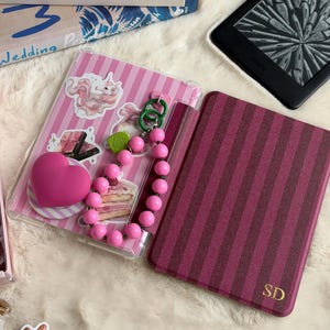 Puede incluir: Una funda protectora para lector electrónico con diseño de rayas rosas y blancas y varias pegatinas, incluyendo un unicornio y un trozo de pastel. Un agarre en forma de corazón rosa y un abalorio de cuentas rosas están unidos a la funda.
