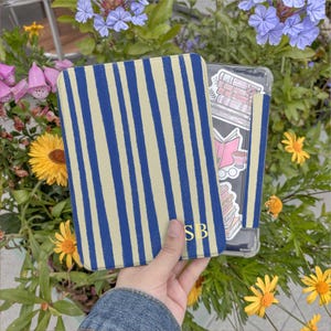 May include: A blue and beige striped notebook cover with the initials "SB" in gold. The notebook is held next to a clear phone case decorated with book-themed stickers. The background features flowers and greenery.