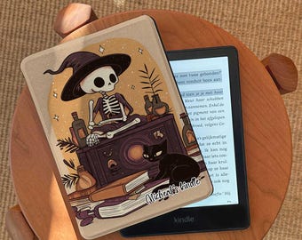 May include: A Kindle e-reader with a decorative cover featuring a skeleton in a witch hat, a black cat, and bottles. The cover is tan with a dark illustration. The Kindle is on a wooden table.