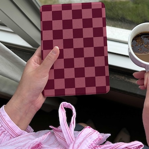May include: A burgundy and light burgundy checkered tablet case. The case is held in a person's hand near a window. A cup of coffee is held in the other hand. The person is wearing a pink striped shirt.