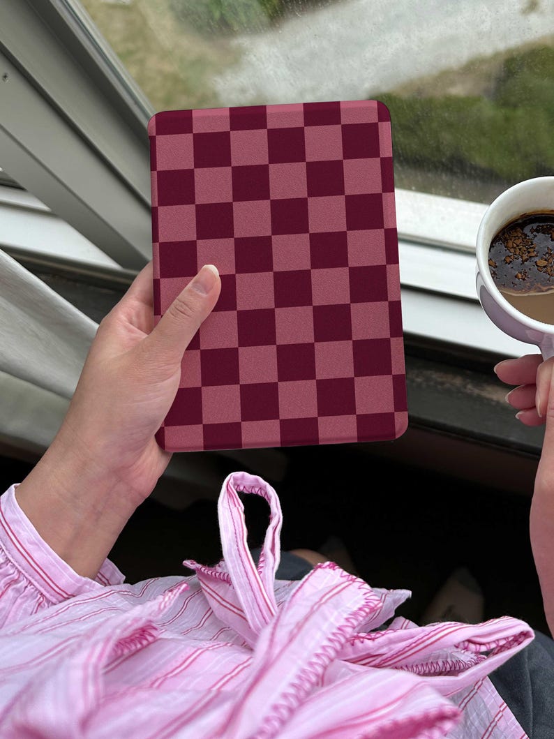 May include: A burgundy and light burgundy checkered tablet case. The case is held in a person's hand near a window. A cup of coffee is held in the other hand. The person is wearing a pink striped shirt.