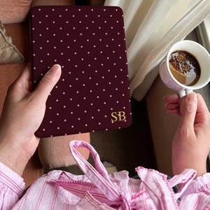May include: A burgundy tablet case with a white polka dot pattern and gold initials "SB". The case is held in a person's hand, with a cup of coffee in the other hand. The person is wearing a pink striped shirt.