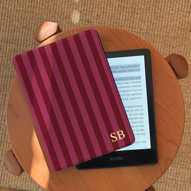 May include: A Kindle e-reader with a burgundy and pink striped cover, displaying text on the screen. The cover has gold initials "SB". The e-reader rests on a small, round wooden table.