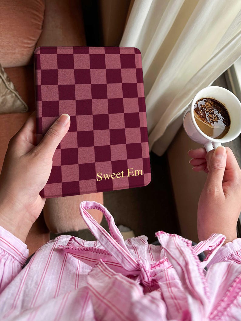 May include: A burgundy and pink checkered tablet case with the text "Sweet Em" in gold lettering. The case is held in a person's hand, with a cup of coffee in the background.