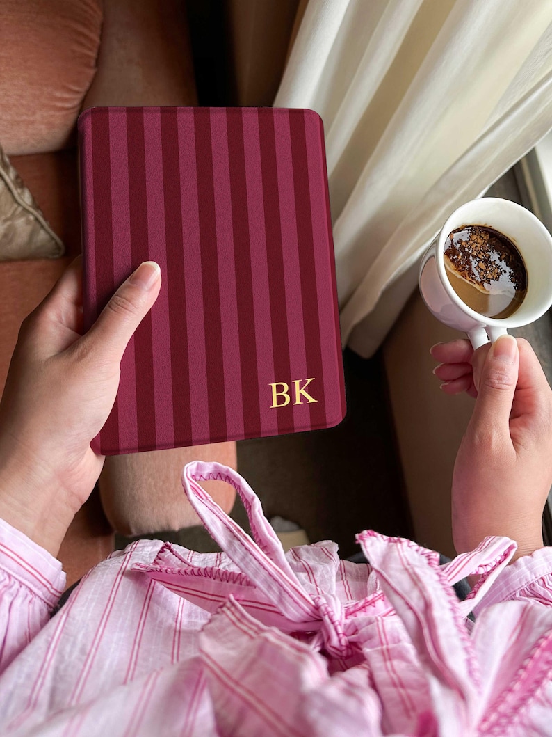 A person holds a Kindle covered in a deep burgundy striped case with gold initials “BK” in the lower corner. The stripes are tonal, giving a subtle classic look. A cup of dark coffee is held in the other hand. Soft morning light filters through cream curtains, and the person is wearing pink striped loungewear, creating a warm, calm reading moment by the window.