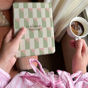 Peut inclure: Une housse de liseuse avec un motif géométrique vert, rose et blanc, avec le texte "I'D RATHER BE Reading". Une tasse blanche de café est tenue dans l'autre main. La personne porte une chemise à rayures roses.