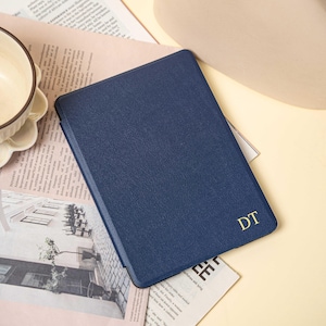 May include: A navy blue tablet case with a textured surface. The initials "DT" are in gold on the lower right corner. The case is resting on a newspaper with a beige background.