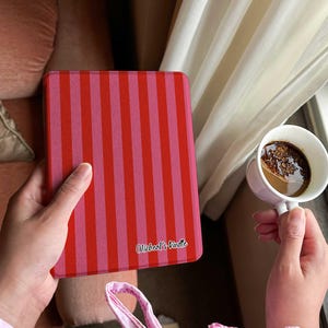Puede incluir: Una funda Kindle a rayas rojas y rosas con el texto "Micheal's Kindle" en cursiva. En la otra mano se sostiene una taza de café blanca con café. La funda se sostiene frente a una ventana con cortinas blancas.