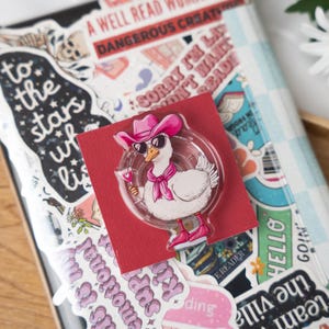 May include: A clear acrylic pin featuring a cartoon goose wearing a pink cowboy hat, sunglasses, scarf, and boots, holding a cocktail. The pin is on a red card, surrounded by stickers and a notebook.