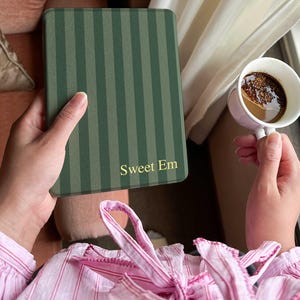 Puede incluir: Una cubierta de libro verde a rayas con el texto "Sweet Em" en letras doradas. Una taza de té blanca con café se sostiene en la otra mano. La persona lleva una camisa a rayas rosas.