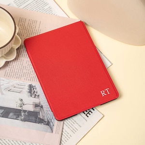 May include: A red tablet case with the initials "RT" in white. The case is rectangular and appears to be made of textured material. The tablet case is on top of a newspaper.