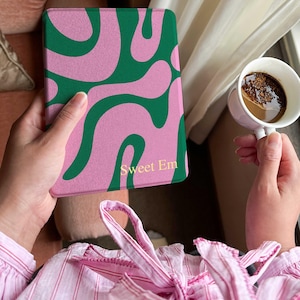 Puede incluir: Una funda rosa para tableta con un diseño abstracto verde y el texto "Sweet Em" en dorado. La funda está sujeta en las manos de una persona, con una taza de café en el fondo.