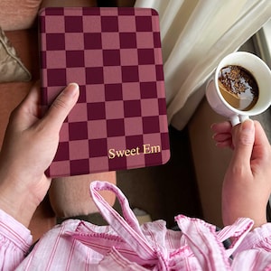 Op de afbeelding: Een bordeauxrode en licht bordeauxrode geruite tablethoes met de tekst "Sweet Em" in gouden letters. De hoes wordt in de handen van een persoon gehouden, met een kopje koffie op de achtergrond.