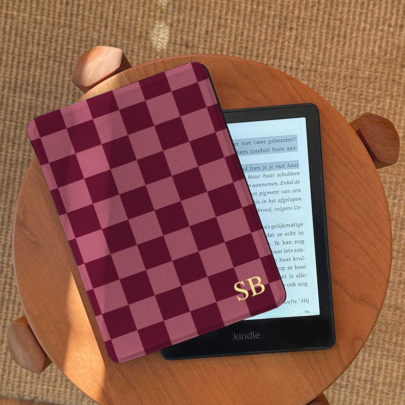 May include: A Kindle e-reader with a burgundy and pink checkered cover, resting on a small wooden table. The cover has gold initials "SB" in the corner. The e-reader displays text on its screen.