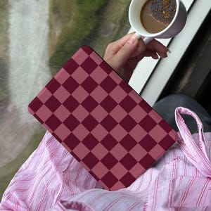 May include: A burgundy and dark pink checkered pattern tablet case rests on a pink striped shirt. A white mug with coffee is held in the background. The scene is set near a window.