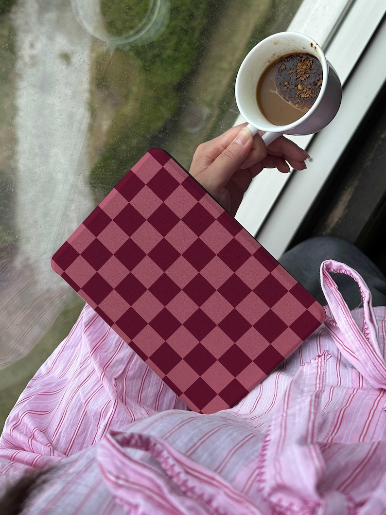 May include: A burgundy and dark pink checkered pattern tablet case rests on a pink striped shirt. A white mug with coffee is held in the background. The scene is set near a window.