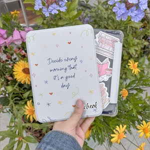 以下が含まれることがあります： 「Decide every morning that it's a good day」という青い文字が書かれた白いタブレットケース。ケースは小さなハート、星、渦巻きで装飾されています。本のステッカーが付いた透明なタブレットケースが後ろに見えます。