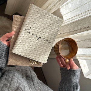 May include: Two light beige woven passport holders, one with the name "Juniper" in cursive and the other with the name "Lyra". A brown mug is held in the hand. The items are set against a neutral background.