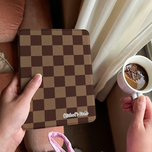 May include: A brown and tan checkered tablet case with the text "Micheal's kindle" is held in a person's hand. A white mug of coffee is held in the other hand. The background includes a pink sofa and a white curtain.