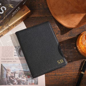 Op de afbeelding: Een zwart leren portemonnee met gouden initialen "SB" ligt op een houten oppervlak. De portemonnee heeft een getextureerde afwerking. Boeken, een hoed, een pen en een glas zijn ook zichtbaar.