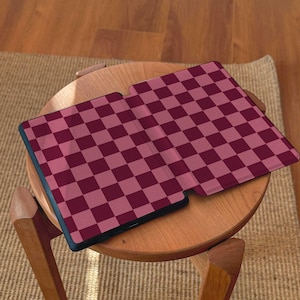 May include: A tablet case with a burgundy and pink checkered pattern. The case is open and rests on a small wooden table. The table has three legs and is positioned on a woven rug.